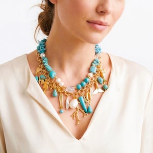 May include: A multi-strand necklace featuring turquoise beads, gold charms, and white pearls. The necklace has a variety of sea-themed charms, including fish, shells, and coral. The necklace is worn over a cream-colored blouse.