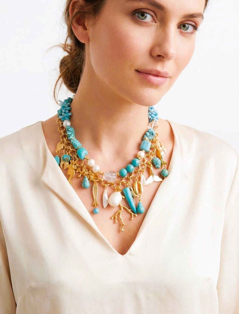 May include: A multi-strand necklace featuring turquoise beads, gold charms, and white pearls. The necklace has a variety of sea-themed charms, including fish, shells, and coral. The necklace is worn over a cream-colored blouse.
