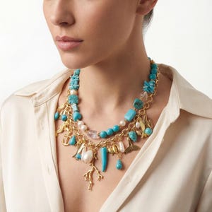 May include: A gold-toned necklace with turquoise, white, and clear beads, and gold charms. The necklace features a variety of shapes, including teardrops, coral, and anchors. The necklace is layered and worn against a cream-colored blouse.