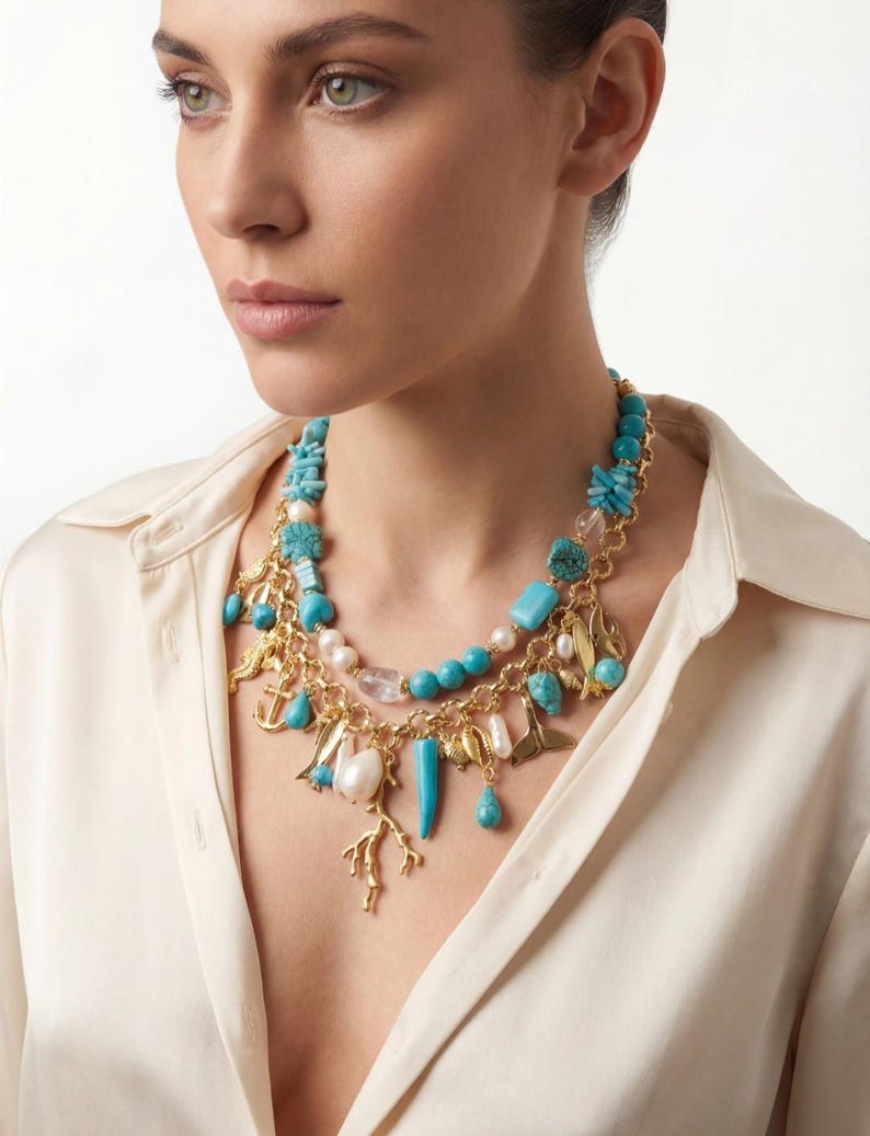 May include: A gold-toned necklace with turquoise, white, and clear beads, and gold charms. The necklace features a variety of shapes, including teardrops, coral, and anchors. The necklace is layered and worn against a cream-colored blouse.