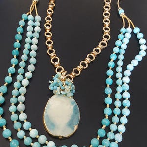 May include: A layered necklace with a large, oval-shaped pendant made of blue and white agate. The pendant is surrounded by smaller blue and white beads. The necklace is made of gold chain and blue beads.