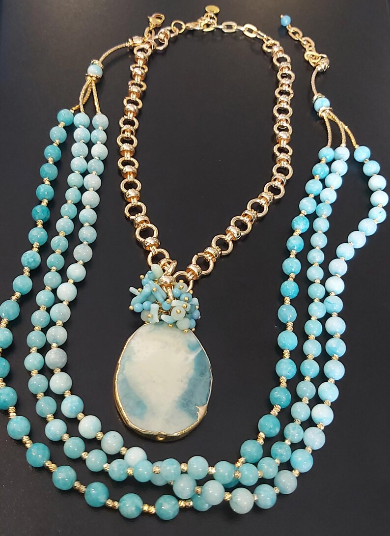 May include: A layered necklace with a large, oval-shaped pendant made of blue and white agate. The pendant is surrounded by smaller blue and white beads. The necklace is made of gold chain and blue beads.