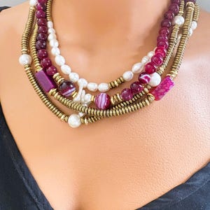 May include: A multi-strand necklace featuring a combination of burgundy beads, white pearls, and gold-toned metal accents. The necklace includes various shapes and sizes of beads, creating a layered effect. The necklace is displayed on a person.