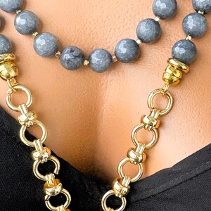 Puede incluir: Un collar de varias hebras con cuentas de piedra gris facetadas intercaladas con peque&ntilde;os espaciadores dorados. El collar contin&uacute;a con una cadena gruesa de tono dorado pulido con eslabones circulares entrelazados que se unen a las secciones.