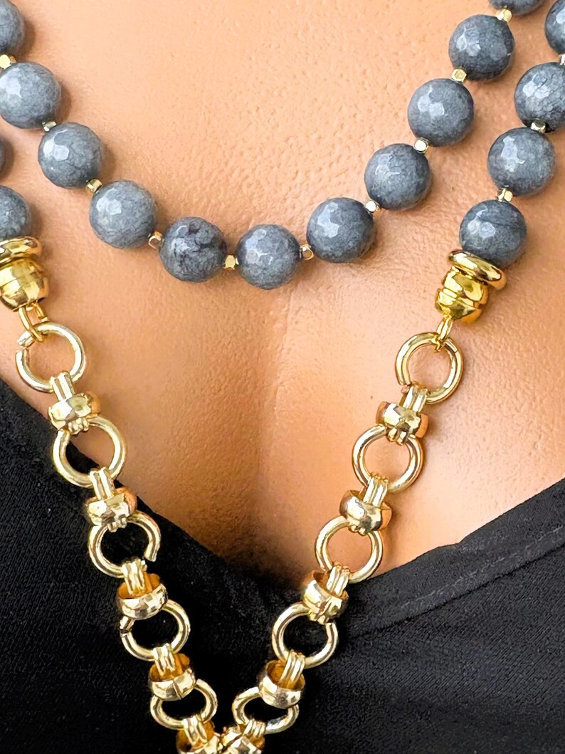 Puede incluir: Un collar de varias hebras con cuentas de piedra gris facetadas intercaladas con peque&ntilde;os espaciadores dorados. El collar contin&uacute;a con una cadena gruesa de tono dorado pulido con eslabones circulares entrelazados que se unen a las secciones.