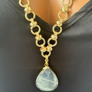 Puede incluir: Un collar de cadena en tono dorado que presenta una serie de eslabones circulares entrelazados. El collar incluye un gran colgante en forma de l&aacute;grima con una piedra lisa de color gris p&aacute;lido engastada en un marco dorado, suspendido del centro.