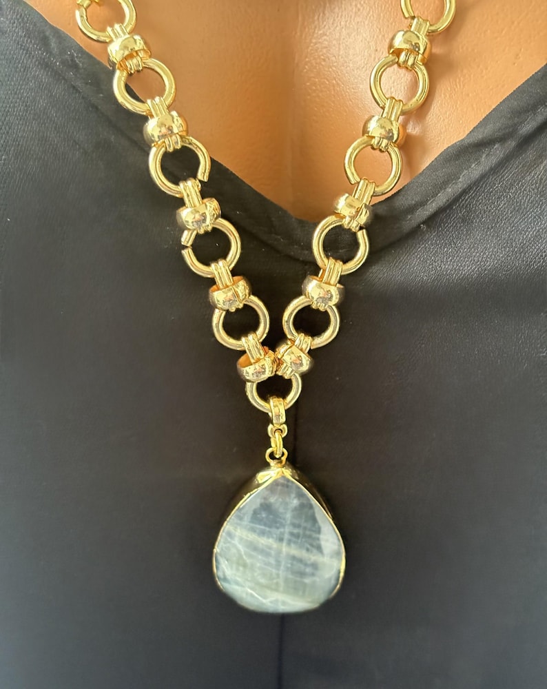 Puede incluir: Un collar de cadena en tono dorado que presenta una serie de eslabones circulares entrelazados. El collar incluye un gran colgante en forma de l&aacute;grima con una piedra lisa de color gris p&aacute;lido engastada en un marco dorado, suspendido del centro.