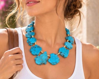 May include: A statement necklace featuring large, irregular, bright turquoise stones with dark matrix veining, interspersed with small, round, metallic gold-toned beads. The necklace rests against a white ribbed tank top.