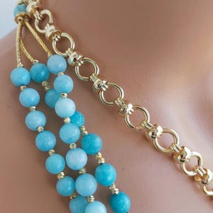 May include: A multi-strand necklace with light blue beads and a gold chain. The necklace is layered with three strands of beads and a single strand of gold chain.