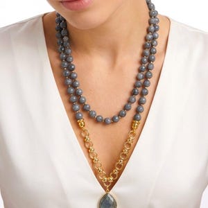 Puede incluir: Un collar de varias hebras con cuentas grises facetadas dispuestas en dos filas, conectadas a una cadena de metal dorado con eslabones circulares. El collar termina en un gran colgante de piedra gris en forma de l&aacute;grima engastado en oro.