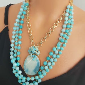 May include: A layered necklace with a large, oval, blue and white agate pendant. The necklace is made of small, round, turquoise blue beads and a gold chain.