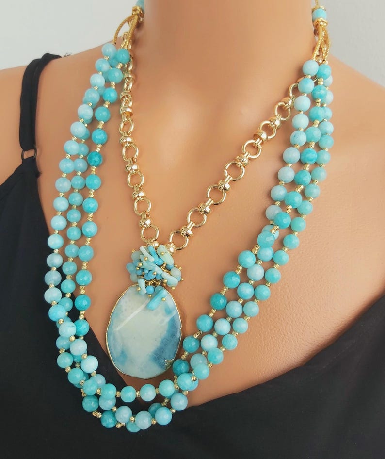 May include: A layered necklace with a large, oval, blue and white agate pendant. The necklace is made of small, round, turquoise blue beads and a gold chain.