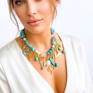 May include: A statement necklace featuring turquoise beads, pearls, and gold-toned charms. The necklace includes seahorse, fish, and coral designs. The necklace is worn over a white top.
