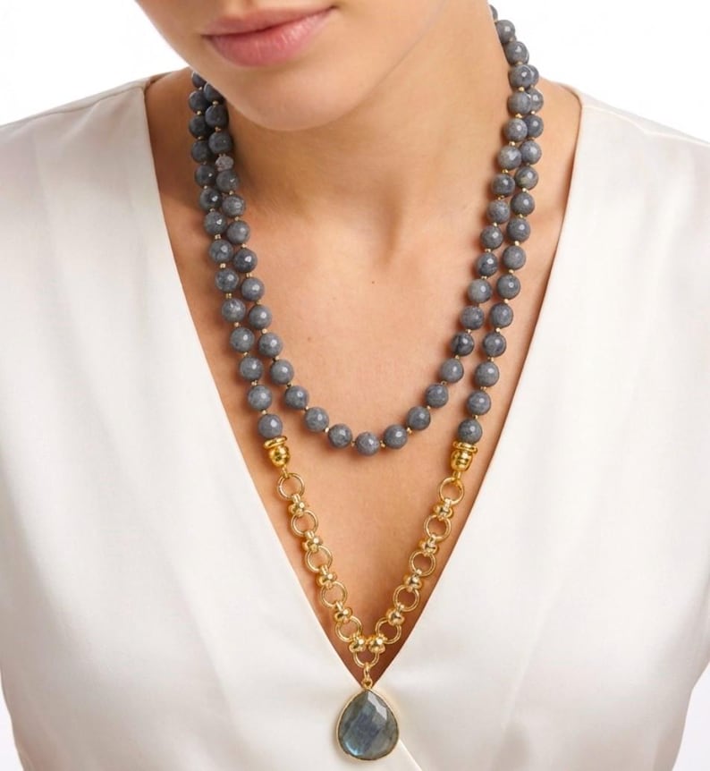 Puede incluir: Un collar de varias hebras con cuentas grises facetadas dispuestas en dos filas, conectadas a una cadena de metal dorado con eslabones circulares. El collar termina en un gran colgante de piedra gris en forma de l&aacute;grima engastado en oro.