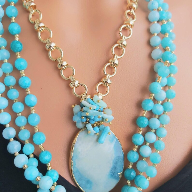 May include: A layered necklace with a large blue agate pendant and smaller blue and white beads. The necklace is made of gold chain and blue beads.