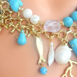 May include: A close-up of a gold-toned necklace with turquoise, white, and pink accents. The necklace features various charms, including a fish, teardrop, and pearl-like beads. The chain has a coral-like design.
