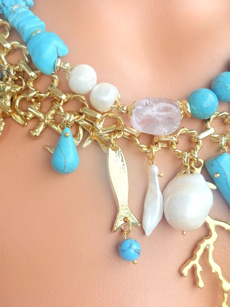 May include: A close-up of a gold-toned necklace with turquoise, white, and pink accents. The necklace features various charms, including a fish, teardrop, and pearl-like beads. The chain has a coral-like design.