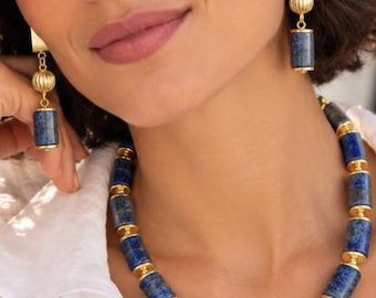 Puede incluir: Un collar y pendientes a juego con cuentas cilíndricas de lapislázuli azul, acentuadas con espaciadores de metal dorado y cuentas esféricas estriadas. El conjunto de joyería muestra un patrón consistente de segmentos de piedra azul profunda separados por herrajes dorados pulidos.