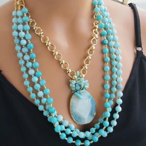 May include: A multi-strand necklace with a large, light blue agate pendant. The necklace is made of small, round, light blue beads and gold chain links. The pendant is surrounded by smaller, light blue stones.