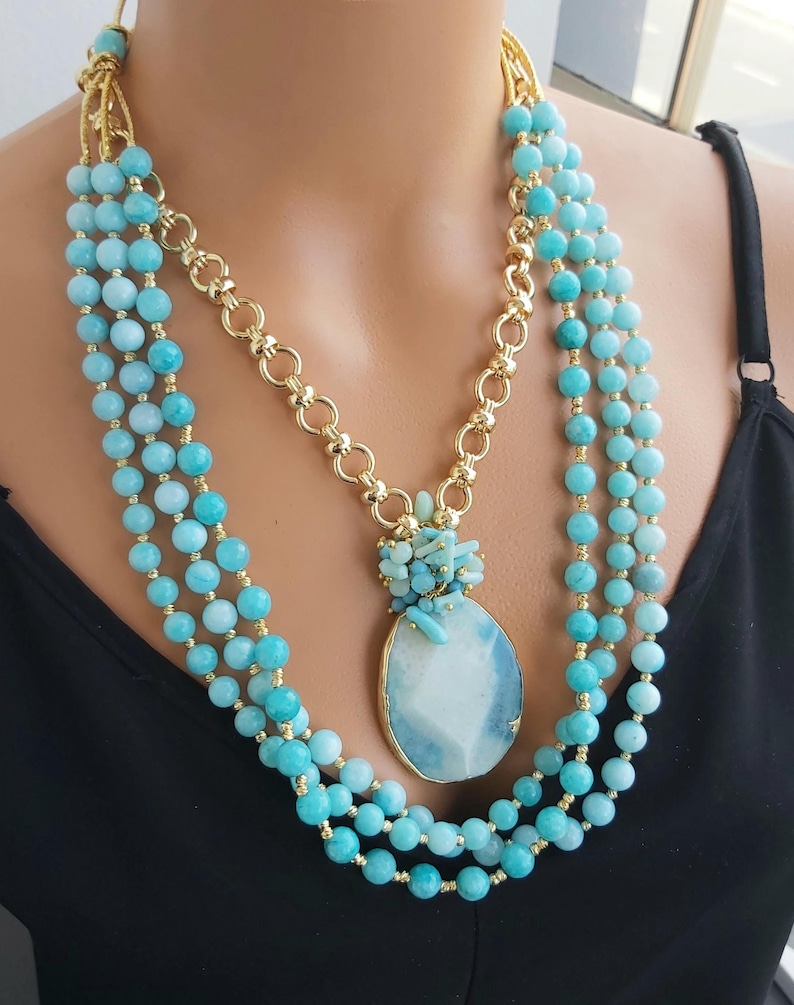 May include: A multi-strand necklace with a large, light blue agate pendant. The necklace is made of small, round, light blue beads and gold chain links. The pendant is surrounded by smaller, light blue stones.