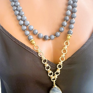 Puede incluir: Un collar de doble hebra con cuentas grises facetadas y una secci&oacute;n de cadena de eslabones dorados. El collar incluye un colgante de piedra gris en forma de l&aacute;grima con un engaste dorado, que descansa sobre una prenda de tela negra.