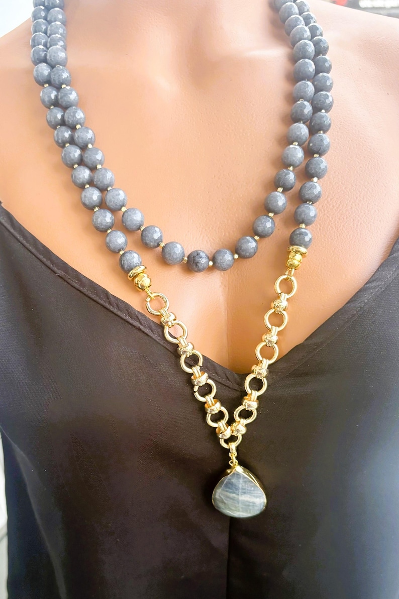 Puede incluir: Un collar de doble hebra con cuentas grises facetadas y una secci&oacute;n de cadena de eslabones dorados. El collar incluye un colgante de piedra gris en forma de l&aacute;grima con un engaste dorado, que descansa sobre una prenda de tela negra.