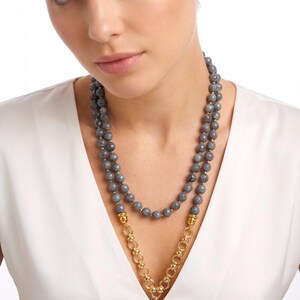 Puede incluir: Un collar de doble hebra con cuentas grises facetadas y una secci&oacute;n de cadena dorada. El collar incluye un colgante central de piedra gris en forma de l&aacute;grima suspendido de un conector de metal dorado, sobre una prenda superior blanca.