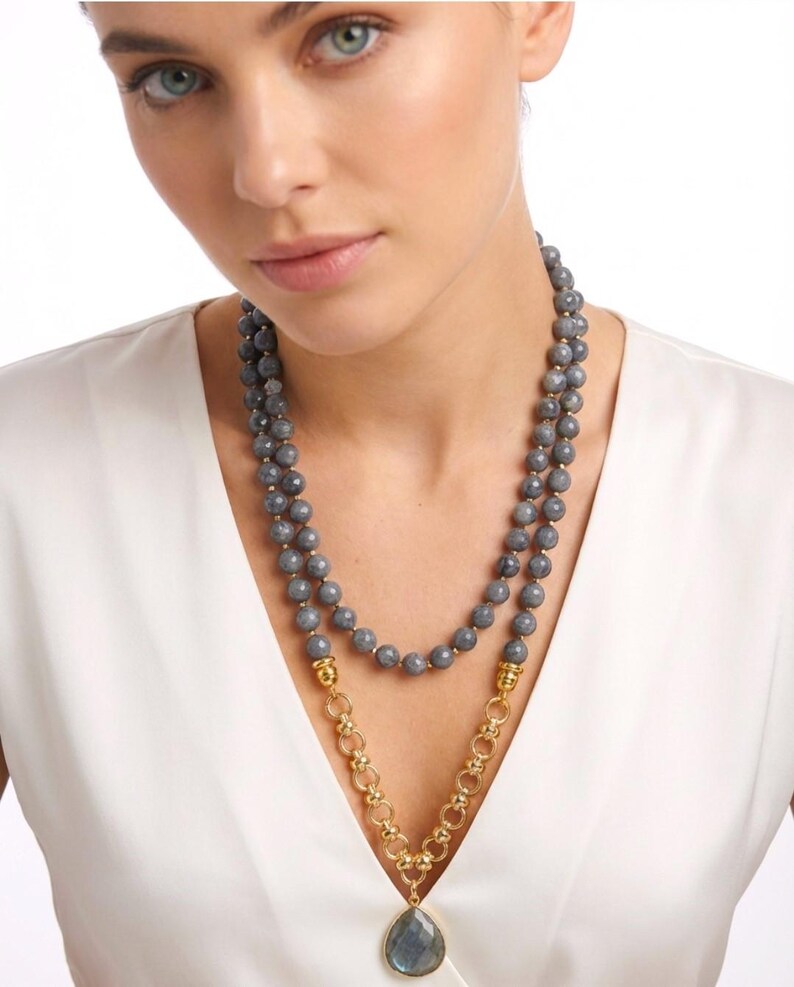Puede incluir: Un collar de doble hebra con cuentas grises facetadas y una secci&oacute;n de cadena dorada. El collar incluye un colgante central de piedra gris en forma de l&aacute;grima suspendido de un conector de metal dorado, sobre una prenda superior blanca.