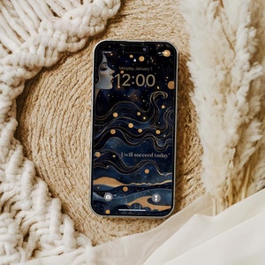 May include: A smartphone shows a celestial wallpaper with a woman's profile, gold details, and the words "I will succeed today." The time is 12:00 on Monday, January 1. The phone is on a textured beige surface.