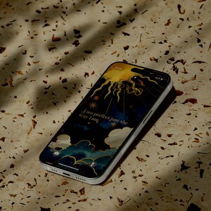 May include: A silver smartphone shows a celestial wallpaper featuring a sun, stars, and clouds. The text "I am perfect just the way I am" is displayed. The phone is on a speckled, light-brown surface, with shadows cast across it.