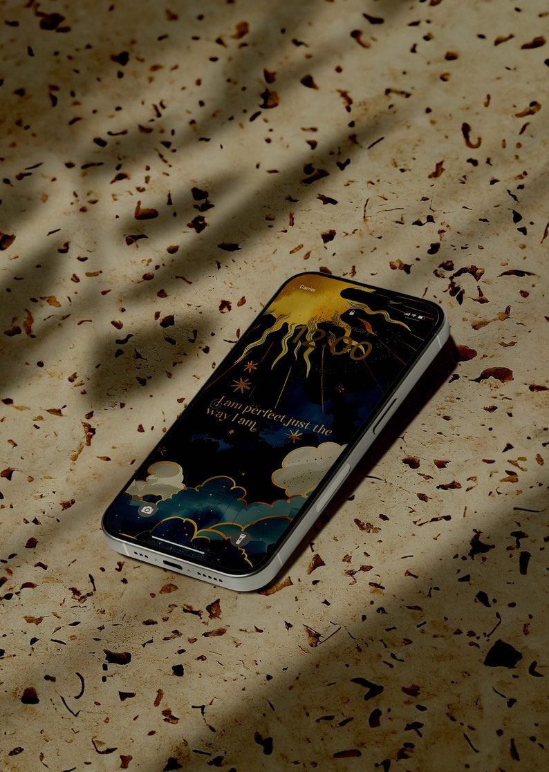 May include: A silver smartphone shows a celestial wallpaper featuring a sun, stars, and clouds. The text "I am perfect just the way I am" is displayed. The phone is on a speckled, light-brown surface, with shadows cast across it.