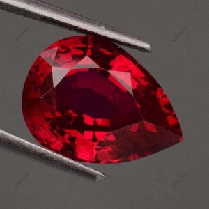 Op de afbeelding: Een gefacetteerde, peervormige robijn edelsteen, vastgehouden door een pincet. De robijn heeft een diepe, verzadigde rode kleur met een hoge mate van helderheid. De facetten van de edelsteen reflecteren licht, waardoor een briljante glans ontstaat. De achtergrond is neutraal grijs.