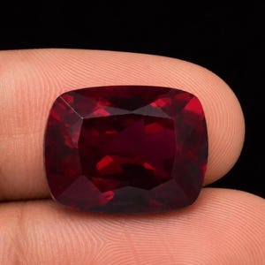 May include: A deep red, cushion-cut gemstone with a faceted surface that reflects light. The stone has a rectangular shape with rounded corners and a rich, saturated crimson hue, displaying internal clarity and sharp, geometric edges throughout.