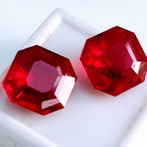May include: Two faceted, octagonal, bright red gemstones. The gems are set against a white background. The gems have a high degree of clarity and reflect light, showcasing their brilliant cut. The overall impression is one of luxury and elegance.