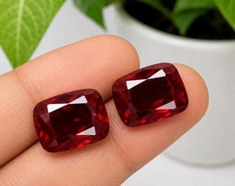 Puede incluir: Dos piedras preciosas de color rojo intenso con talla cojín y superficie facetada que refleja la luz. Las piedras tienen forma rectangular con esquinas redondeadas, mostrando un tono carmesí rico y saturado en su estructura cristalina pulida.