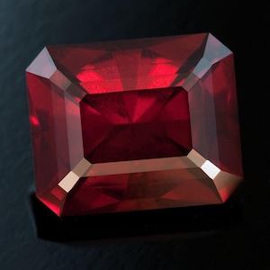 May include: A faceted, rectangular-cut, deep red gemstone. The gem has a polished surface with sharp edges and a reflective interior. The gemstone is set against a dark background, highlighting its color and clarity. The gem's shape is a rectangle with cut corners.