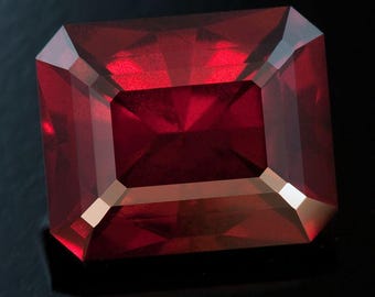 Puede incluir: Una gema roja oscura, tallada en forma rectangular facetada. La gema tiene una superficie pulida con bordes afilados y un interior reflectante. La piedra preciosa se encuentra sobre un fondo oscuro, resaltando su color y claridad. La forma de la gema es un rectángulo con esquinas cortadas.