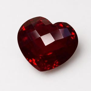 May include: A deep red, heart-shaped gemstone with a faceted surface. The stone features multiple geometric cuts that create light reflections and shadows across its dark crimson surface, highlighting the intricate, polished structure of the gem.