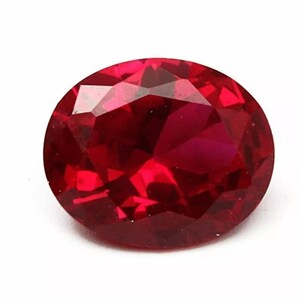 Peut inclure: Une pierre précieuse de forme ovale, un rubis facetté. La gemme est d'un rouge profond et saturé, avec de multiples facettes qui réfléchissent la lumière. La pierre est posée sur un fond blanc.