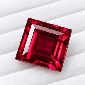 May include: A square-cut, vibrant red gemstone with faceted surfaces that create sharp geometric reflections. The stone displays a deep, saturated crimson hue with clear, transparent clarity and a polished finish, showcasing a precise, symmetrical shape.