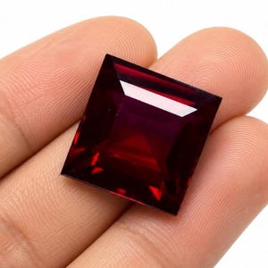 May include: A square-cut, deep red gemstone with a faceted surface that reflects light. The stone features sharp, clean edges and a translucent, dark crimson color throughout, showcasing a geometric shape with a polished, smooth finish.