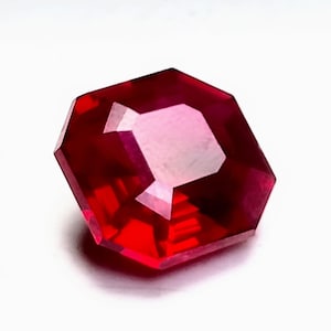 May include: A faceted, octagonal-cut ruby gemstone. The gem is a deep, saturated red color with a high degree of clarity. The facets reflect light, creating a brilliant sparkle. The gemstone is set against a white background.