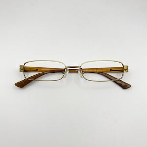 May include: Gold-toned rectangular eyeglasses with brown temples and a nose bridge. The glasses have a classic design and are set against a white background. The frame is made of stainless steel.