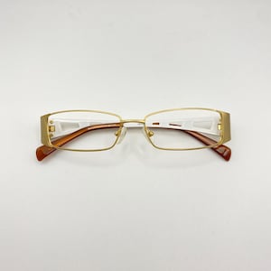 Op de afbeelding: Goudkleurige bril met rechthoekige glazen. De monturen hebben een gouden afwerking met witte en bruine accenten op de slapen en armen. De bril is ontworpen voor gezichtscorrectie en staat op een witte achtergrond.