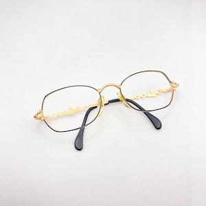 Può includere: Occhiali in metallo dorato con montatura nera e un dettaglio a catena decorativo sul ponte.