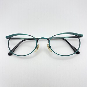 Może przedstawiać: Turkusowe okulary z cienką metalową oprawką i okrągłymi soczewkami. Oprawka ma unikalny design z małymi otworami wzdłuż górnej krawędzi. Tekst "NEWWINE 1076 52[]21 []302" jest widoczny na oprawce. Wyprodukowano we Włoszech.