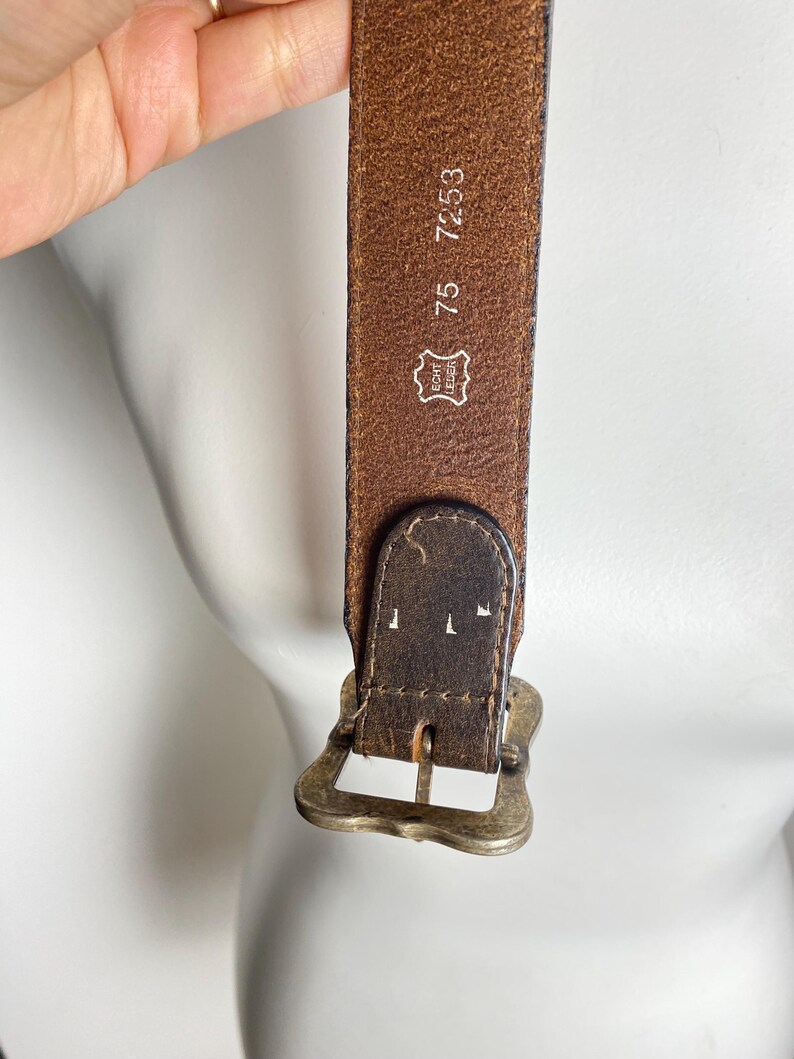 May include: A brown leather belt featuring a weathered texture and a brass-toned metal buckle. The inner side of the belt displays the embossed text 75, 7253, and a stamp reading Echt Leder, indicating genuine leather construction.