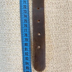 May include: A dark brown belt with four visible adjustment holes positioned next to a blue measuring tape. The tape shows the belt section spans from approximately 72 to 85 centimeters, indicating the length of this specific leather belt segment.
