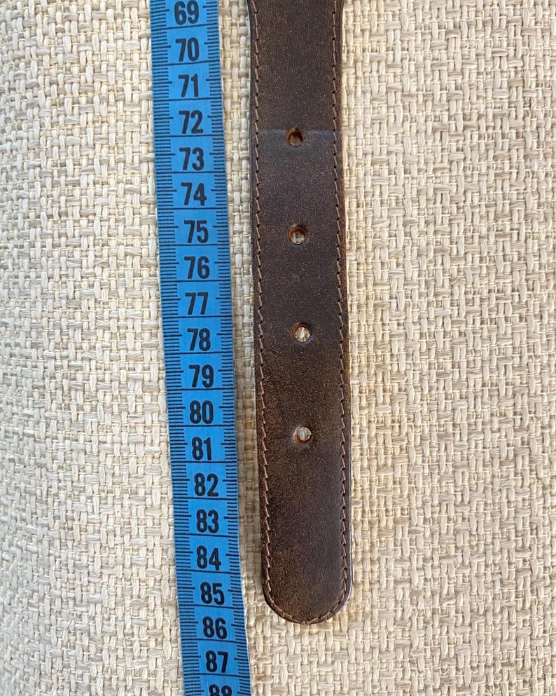 May include: A dark brown belt with four visible adjustment holes positioned next to a blue measuring tape. The tape shows the belt section spans from approximately 72 to 85 centimeters, indicating the length of this specific leather belt segment.