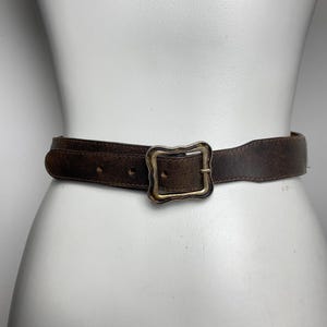 May include: A dark brown leather belt featuring a weathered, distressed texture and a rectangular brass-toned metal buckle with a wavy, decorative edge. The belt has visible stitching along the edges and multiple punched holes for adjustment.